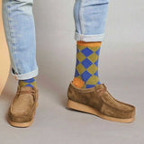 A man wearing a pair of socks featuring a green and blue diamond pattern, in jeans and brown shoes