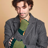 A man wearing a dark gray wool coat and a pair of green and yellow striped fingerless knit gloves, posed against a solid beige background.