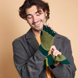 A man wearing a gray wool coat over a yellow shirt models a pair of green and yellow striped fingerless gloves against a solid beige background.