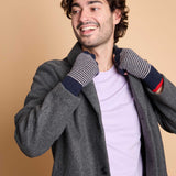 A man wearing a gray wool coat over a light purple t-shirt, featuring striped knit winter gloves with navy and red cuffs, posed against a solid beige background.