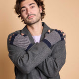 A man wearing a gray knit cardigan and striped fingerless gloves with navy blue fingertips, posed with his arms crossed against a solid beige background.
