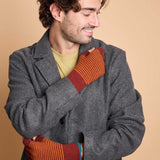 A man wearing a gray wool-blend coat over a yellow-green t-shirt, holding his arms close to his chest to show a pair of orange and red striped knit mittens against a solid beige background.