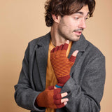 A man wearing a gray coat over a mustard yellow shirt, holding up a pair of fingerless knit gloves with rust red and orange horizontal stripes against a solid beige background.