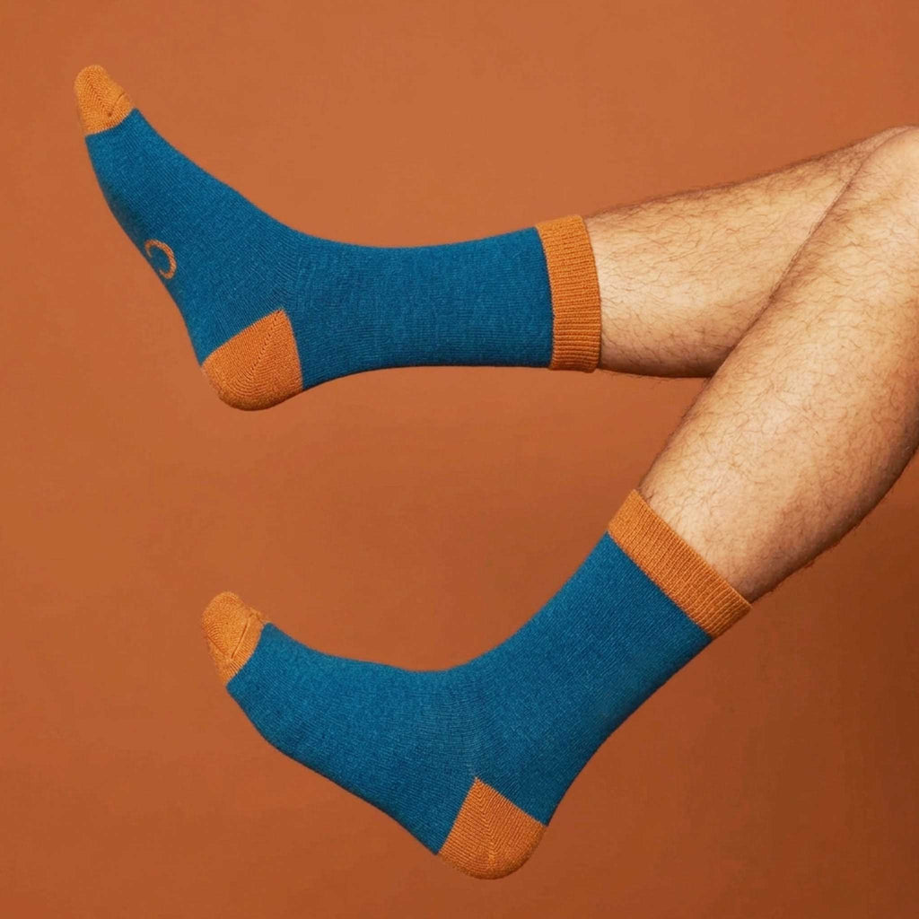 A PAIR OF TEAL WOOL ANKLE SOCKS WITH ORANGE TRIM MODELLED BY A MAN ON AN ORANGE BACKGROUND