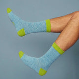 A PAIR OF JADE WOOL ANKLE SOCKS WITH BRIGHT GREEN TRIM MODELLED BY A MAN ON A BLUE BACKGROUND