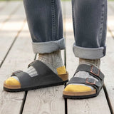 Person wearing black sandals with yellow soles and rolled-up jeans on a wooden floor.