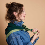 A woman in profile wearing a green knit scarf with a light green stripe over a blue denim shirt, set against a solid beige background.