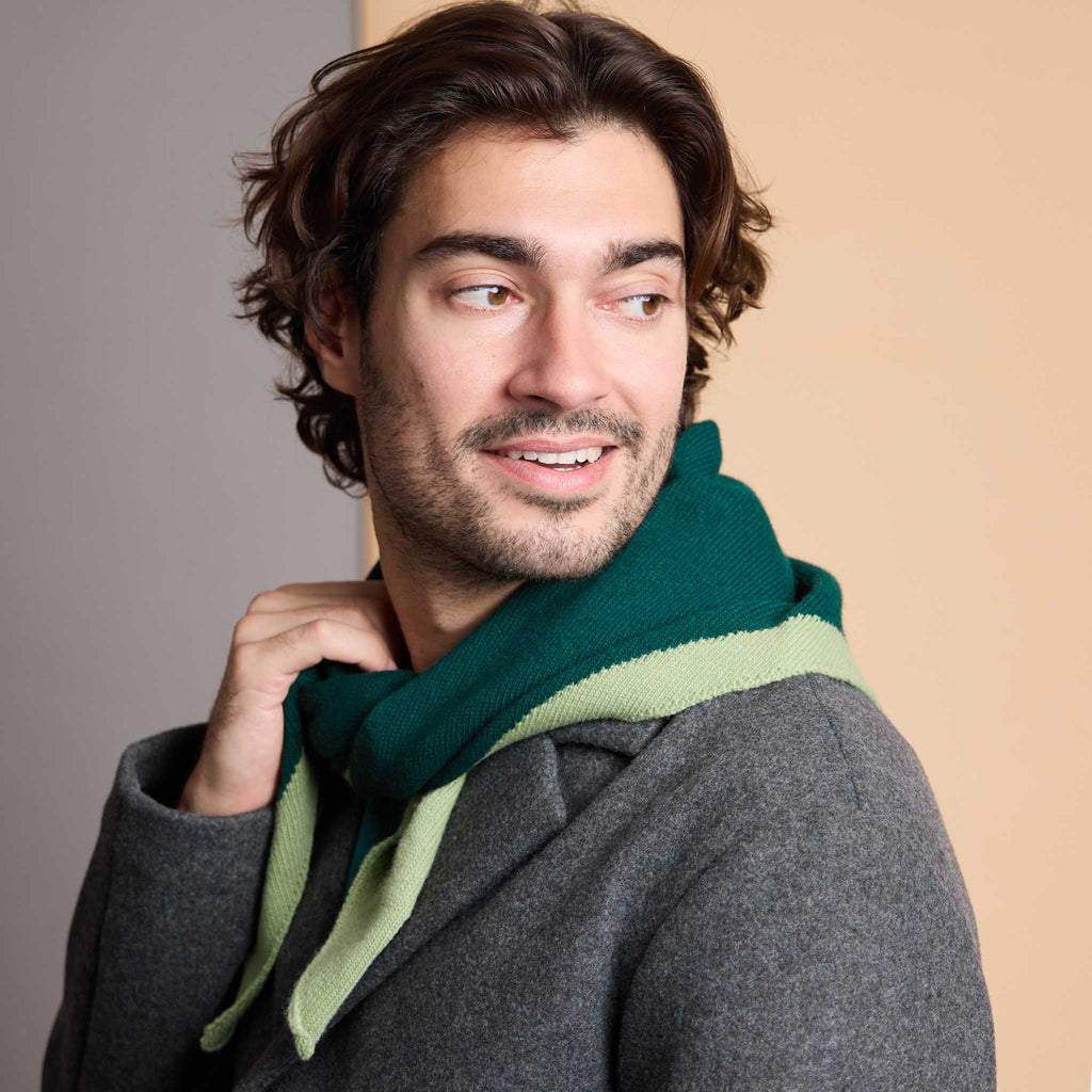A man wearing a dark gray coat and a knit scarf in dark green and light sage green, posed against a split beige and gray background.
