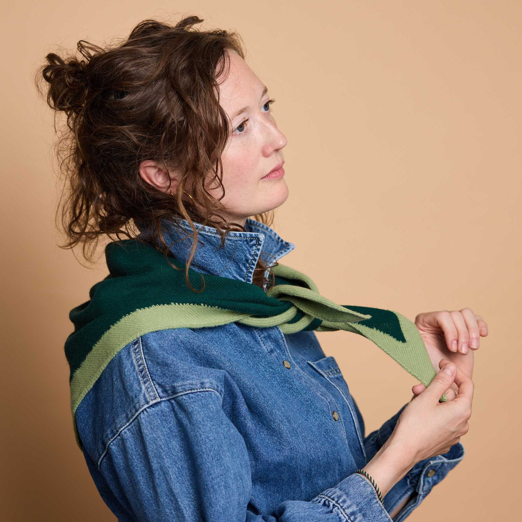 A woman with curly brown hair wearing a blue denim shirt and a green knit scarf, posed against a solid beige background.