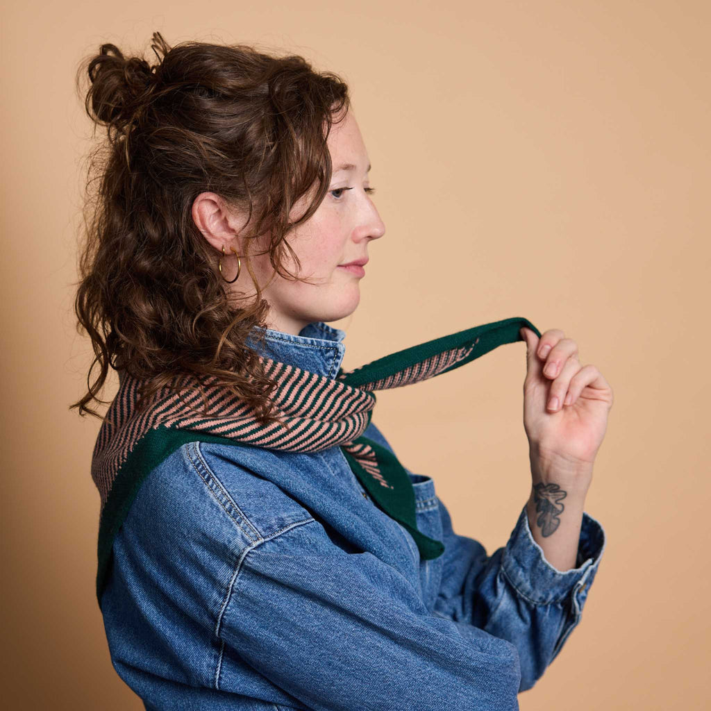 A woman with curly brown hair is shown in profile wearing a blue denim shirt and a striped scarf with a dark green border, holding the scarf's ends against a solid beige background.