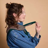A woman with curly brown hair is shown in profile wearing a blue denim shirt and a striped scarf with a dark green border, holding the scarf's ends against a solid beige background.