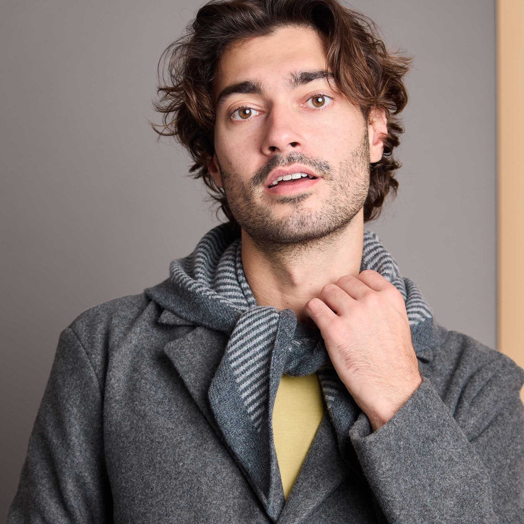 A man wearing a gray wool coat and a gray-and-white striped knit scarf, with a yellow shirt visible underneath, photographed against a plain gray background.