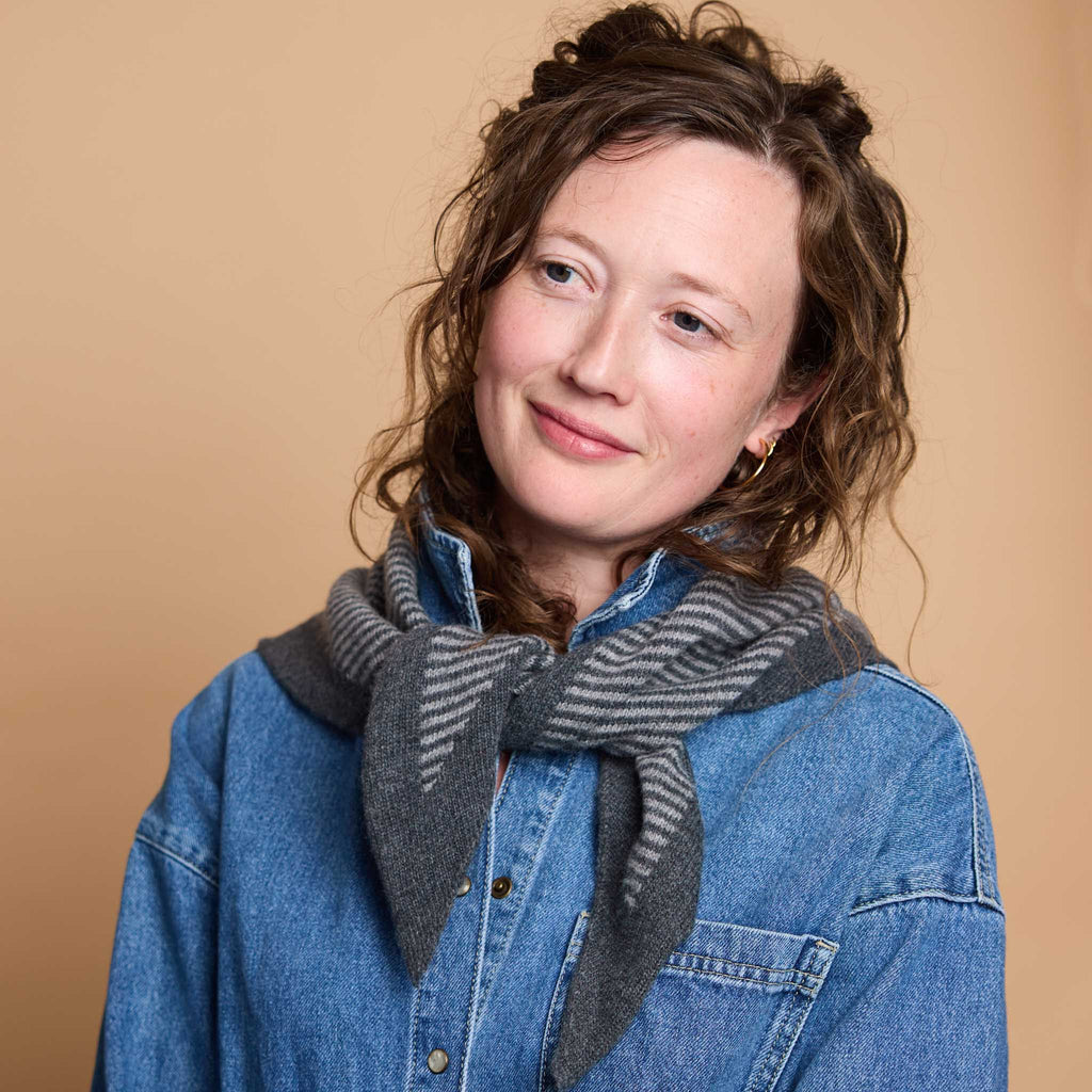 A woman with curly brown hair wearing a blue denim shirt and a gray striped knit scarf, photographed against a solid warm beige background.