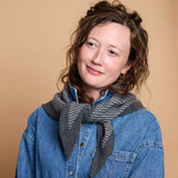 A woman with curly brown hair wearing a blue denim shirt and a gray striped knit scarf, photographed against a solid warm beige background.