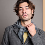 A man wearing a gray wool coat and a gray-and-white striped knit scarf, with a yellow shirt visible underneath, photographed against a plain gray background.