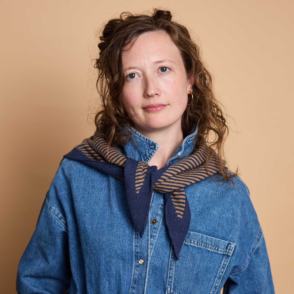 A woman with curly brown hair wearing a blue denim shirt and a navy blue scarf with brown stripes, posed against a solid beige background.