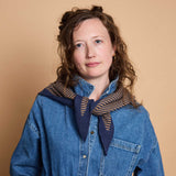 A woman with curly brown hair wearing a blue denim shirt and a navy blue scarf with brown stripes, posed against a solid beige background.