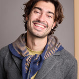 A smiling man with wavy brown hair and light stubble, wearing a gray coat, a blue and tan striped knit scarf, and a yellow shirt, photographed against a plain gray wall.
