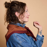 A woman in profile wearing a rust-orange and burgundy striped knit scarf with a solid burgundy border, styled over a blue denim jacket against a solid beige background.