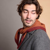 A man wearing a gray blazer and a striped knit scarf in rust orange and burgundy, photographed against a soft beige studio background.