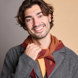 A smiling man with wavy brown hair and a light beard, wearing a gray wool-blend jacket over a mustard yellow shirt and a rust-red and orange striped knit scarf, posed against a split beige and gray studio background.