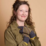 A woman with curly brown hair wearing an olive green jacket and gray knit gloves with yellow stripes, posing against a solid beige background.
