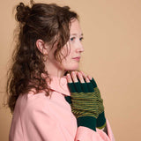 A woman in a pink shirt wearing dark green knit fingerless gloves with yellow stripes, posed against a solid beige background.