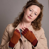 A woman wearing a beige quilted jacket and striped knit fingerless gloves in rust orange and dark red, posing against a neutral gray background.
