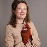 A woman with curly brown hair wearing a beige quilted jacket and rust-orange striped fingerless knit gloves, posing against a solid gray studio background.
